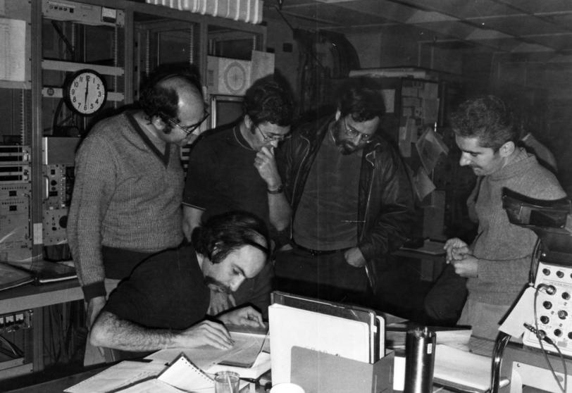 Four men looking down at a fifth who is seated at a table, writing